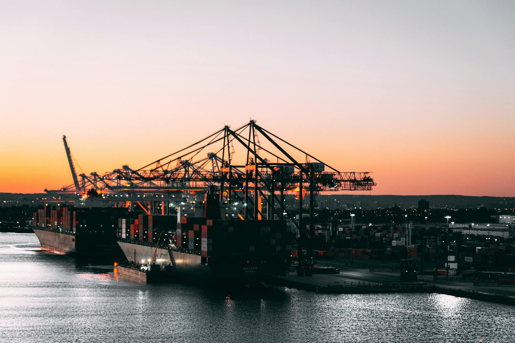 Cargo ships and cranes operating at a commercial harbor during sunset.
